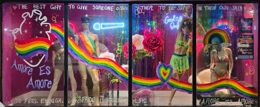 Crave Romance Boutique Pride Month window display featuring rainbow art, mannequins in lingerie, neon signs, and hand-painted text reading "Amore Es Amore" and "Pride Is Freedom" on a hot pink background in Lafayette, Louisiana